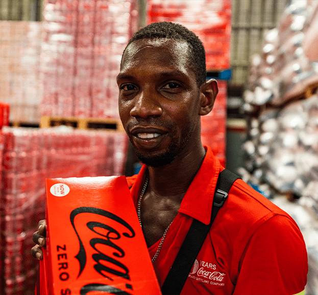 A man with a coca cola shirt holding a box of coca cola with in the background pallets full of coca cola