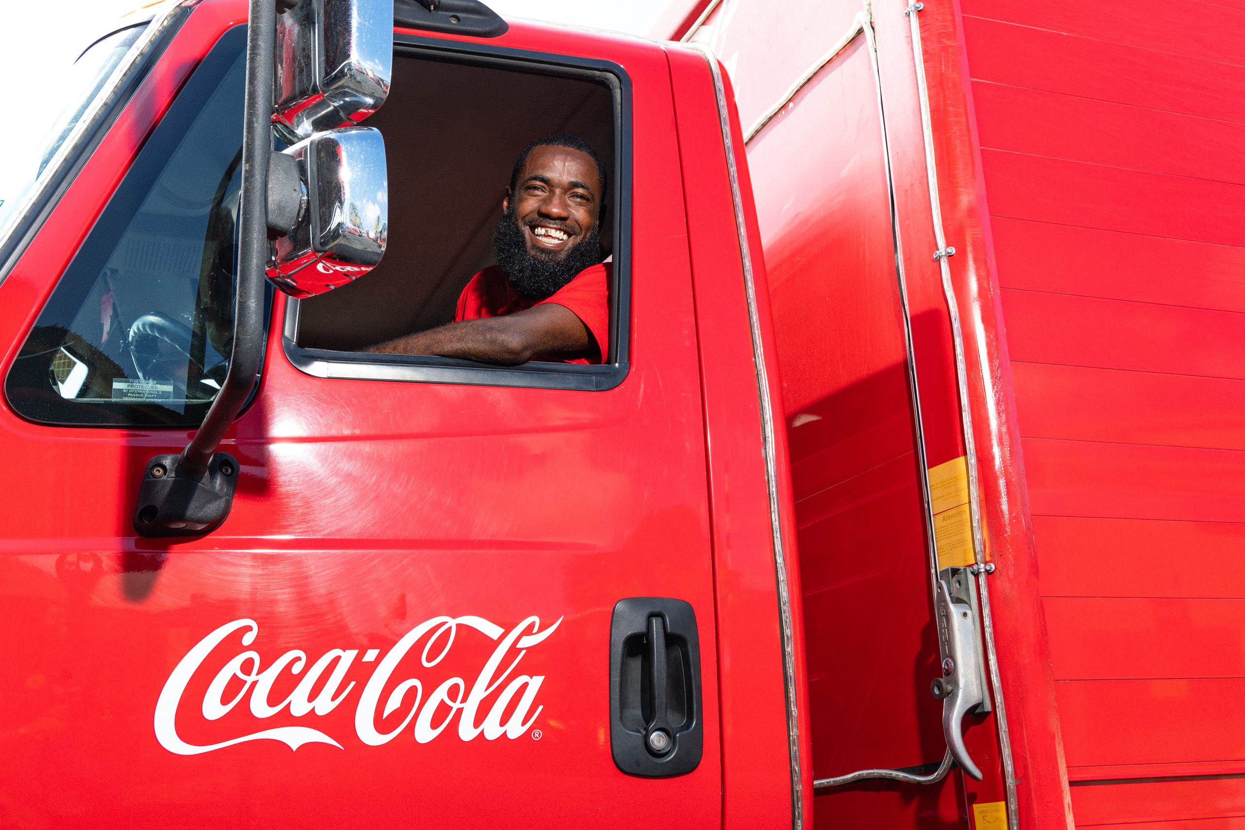 Coca-Cola delivery driver smiling from the window of his red truck, representing commitment and service.