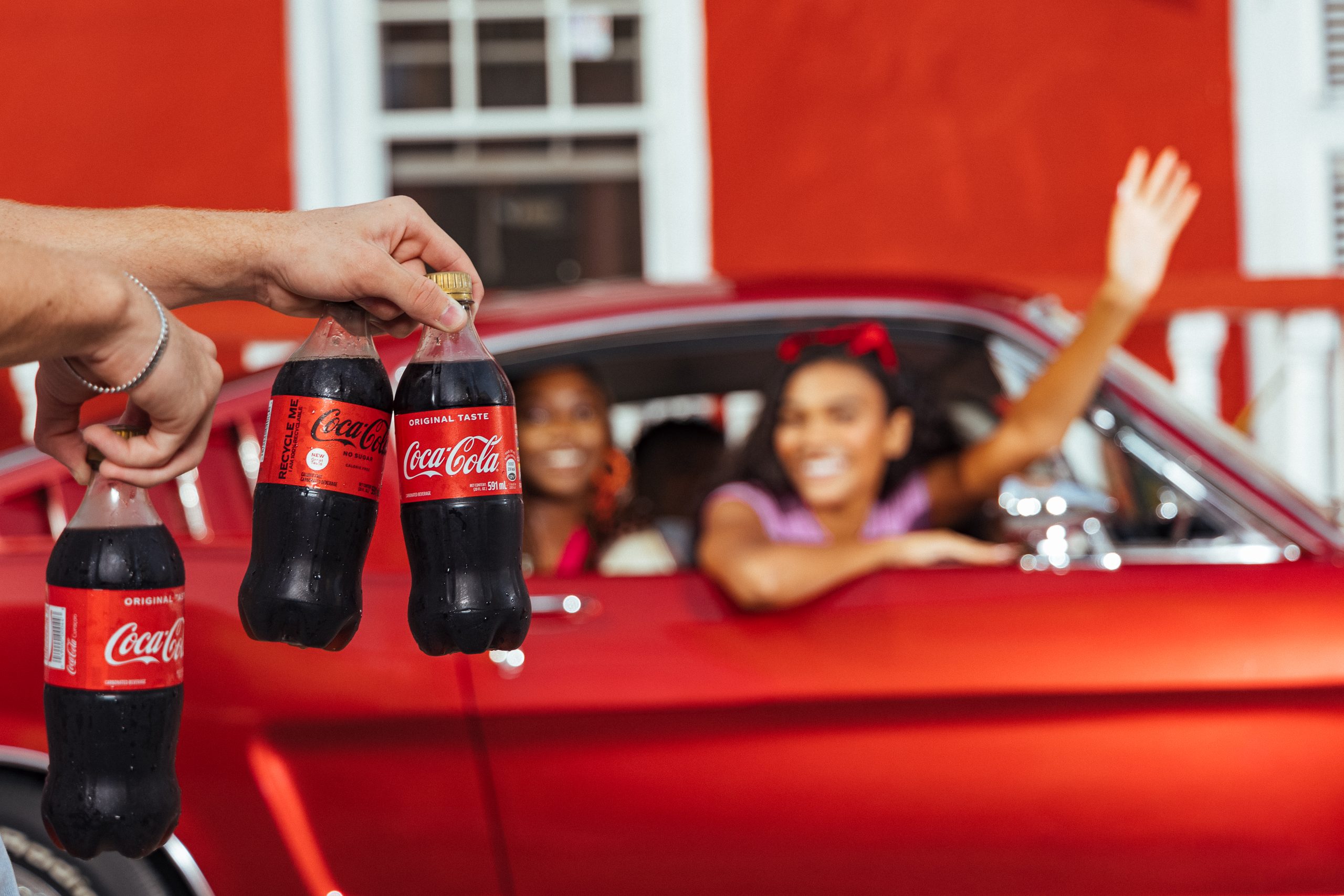 Hand holding three glass Coca-Cola bottles in front of a red classic car with people smiling inside.